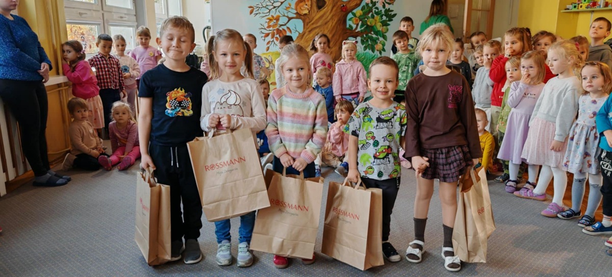 Dzieci, które otrzymały w konkursie nagrody pozują do wspólnego zdjęcia. W tle pozostałe dzieci z przedszkola. Ujęcie 1