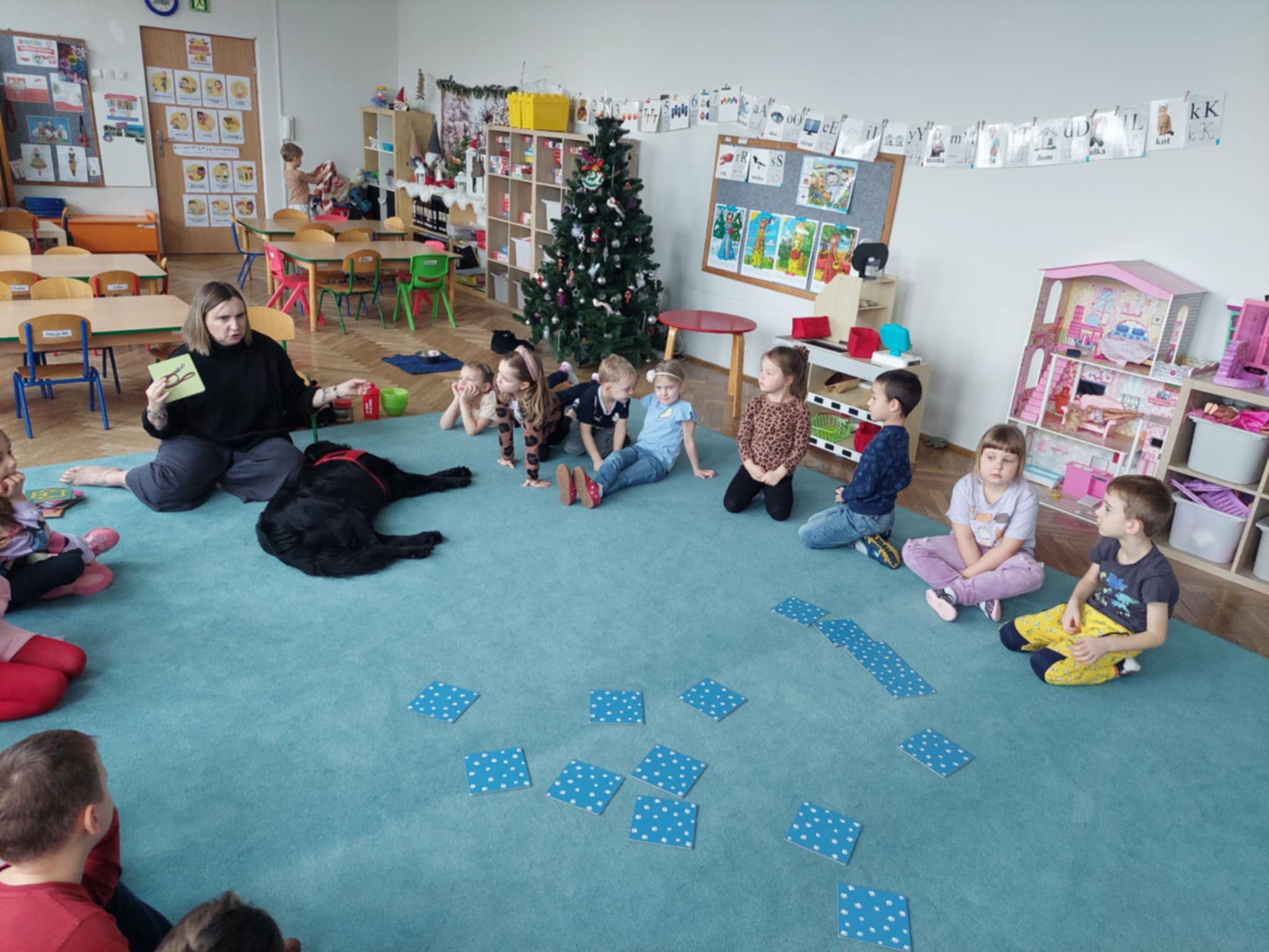Dzieci i nauczycielka siedzą na niebieskim dywanie w sali przedszkolnej. Na zdjęciu widoczny jest duży, czarny pies. Pomiędzy dziećmi leżą w rozsypce karty do gry w "Menory". Ujęcie 1