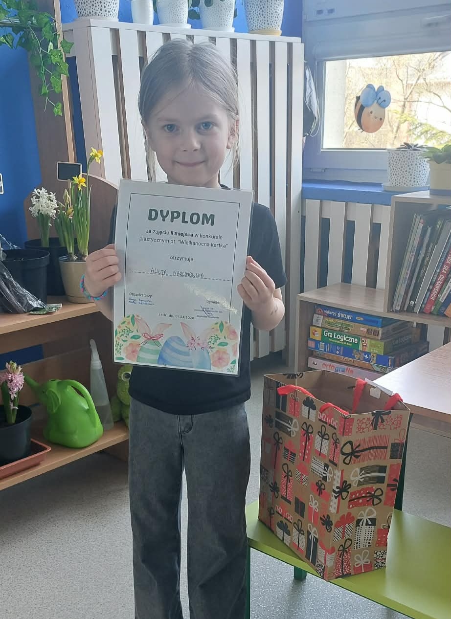Dziecko trzyma pamiątkowy dyplom, a obok stoi torba z nagrodami.