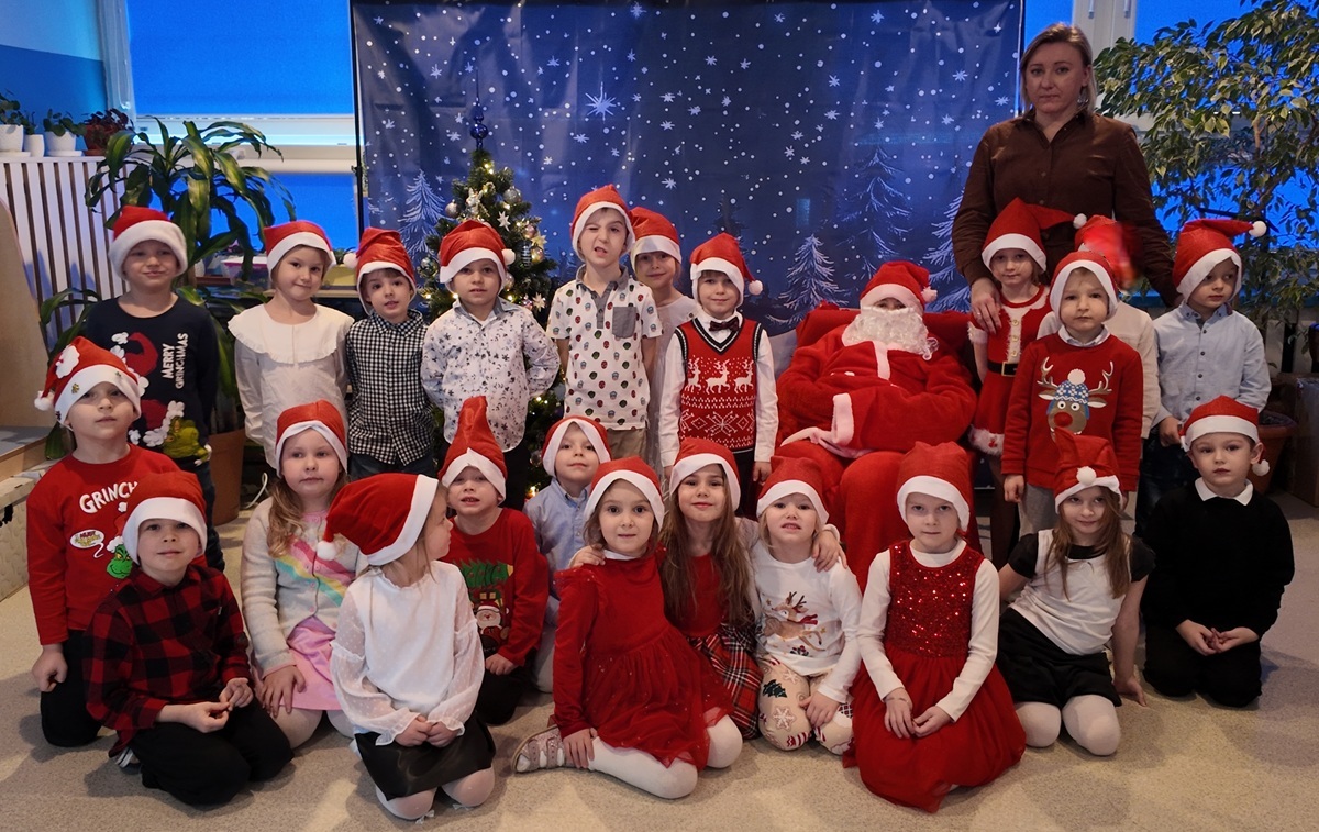 Grupa dzieci i nauczycielka pozują do wsp&oacute;lnego zdjęcia z Mikołajem. Dzieci mają na głowach charakterystyczne czapki. Mikołaj siedzi na fotelu. Zdjęcie częściowo anonimizowane.