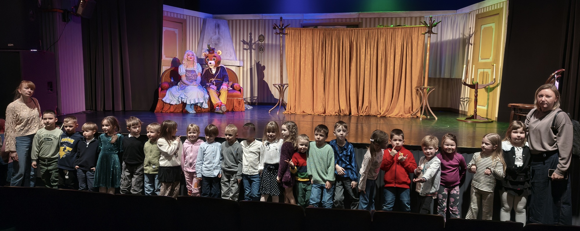 Dzieci i nauczycielki pozują do wsp&oacute;lnego zdjęcia na tle sceny i aktor&oacute;w. Grupa 3