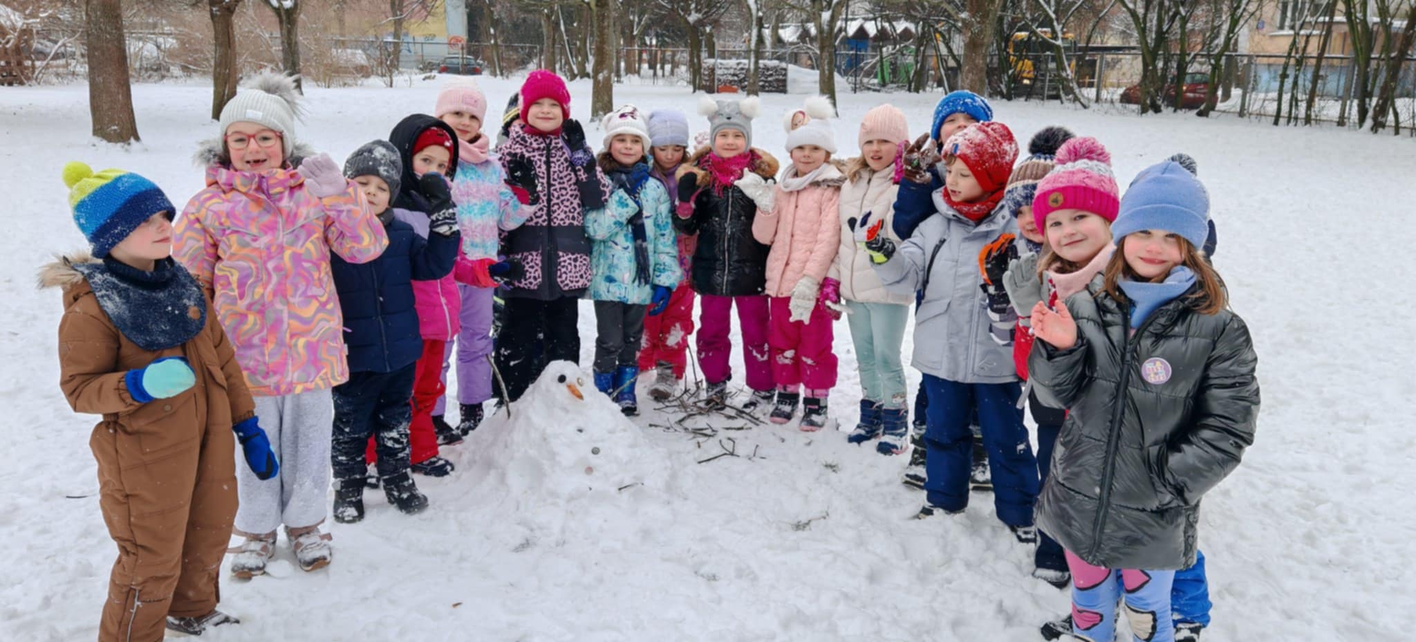 Grupa dzieci pozuje do wsp&oacute;lnego zdjęcia w zimowej scenerii ogrodu przedszkolnego.