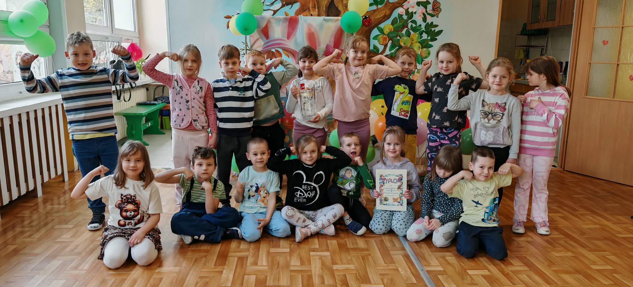 Dzieci z grupy 4 pozują do wsp&oacute;lnego zdjęcia w przedszkolu trzymając pamiątkowy dyplom i puchar.