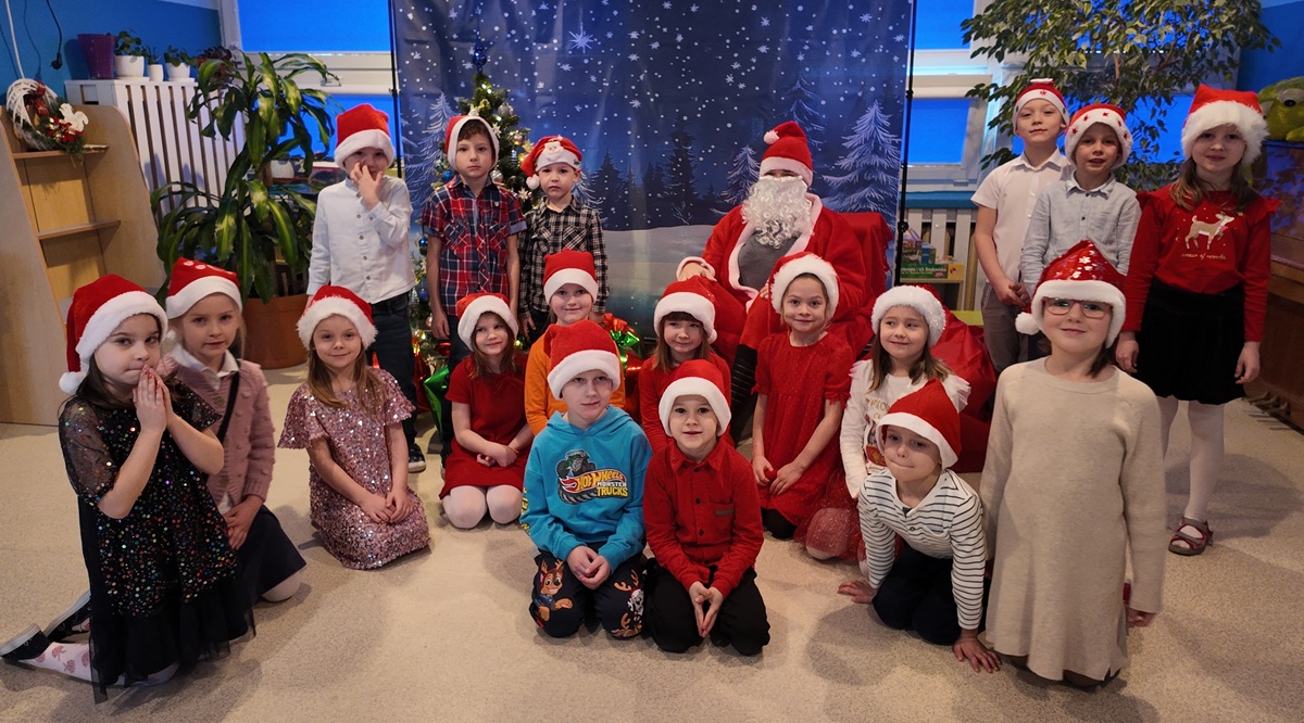 Grupa dzieci pozuje do wsp&oacute;lnego zdjęcia z Mikołajem. Dzieci mają na głowach charakterystyczne czapki. Mikołaj siedzi na fotelu.