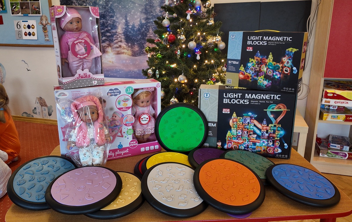 Prezenty jakie grupa 4 dostała od Mikołaja: lalki, klocki i dyski sensoryczne. W tle ubrana choinka i sala przedszkolna.