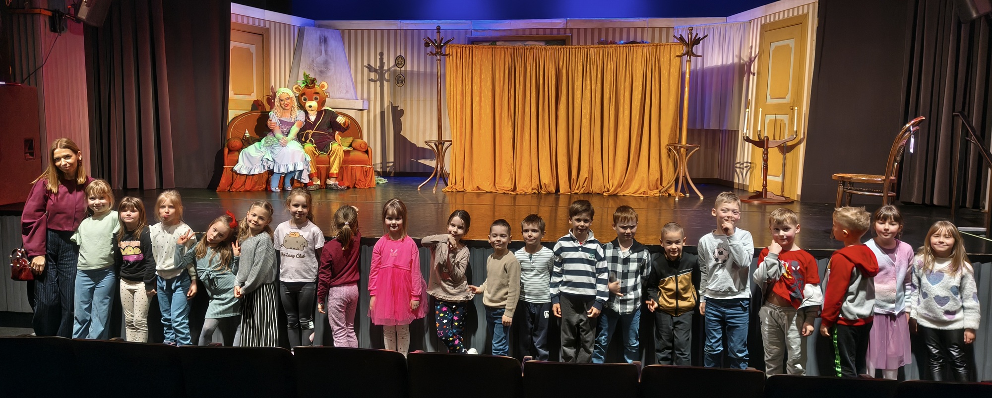 Dzieci i nauczycielka pozują do wsp&oacute;lnego zdjęcia na tle sceny i aktor&oacute;w. Grupa 4