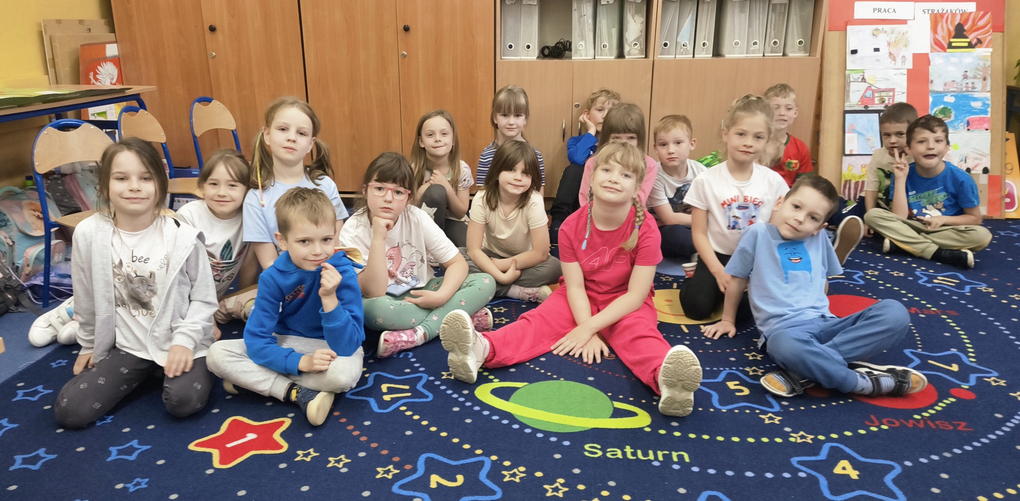 Dzieci siedzą na niebieskim dywanie w sali szkolnej i czekają na rozpoczęcie zajęć. 