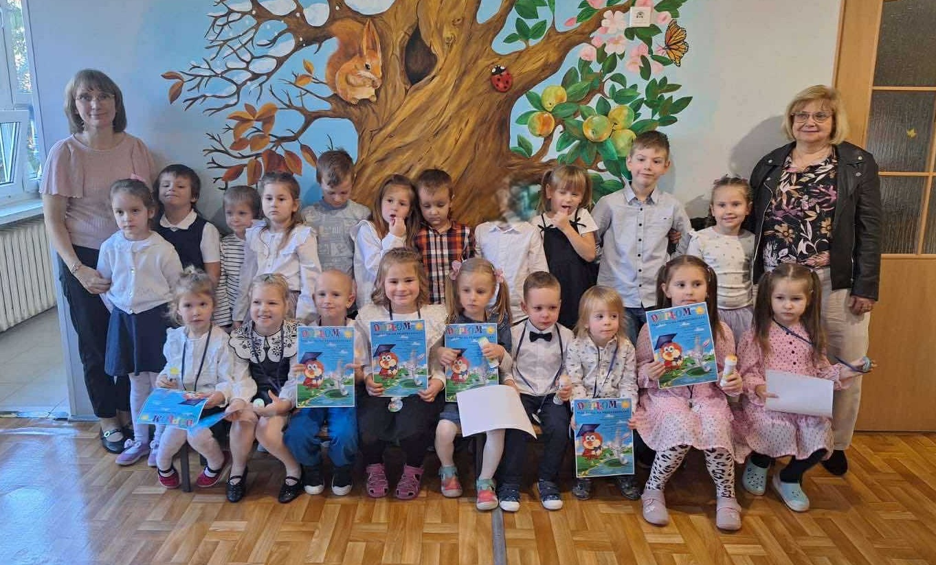 Dzieci i nauczycielki pozują do wspólnego zdjęcia z okazji "Pasowania na przedszkolaka" młodszych kolegów. Dzieci trzymają w rękach pamiątkowe dyplomy. Zdjęcie częściowo anonimizowane.