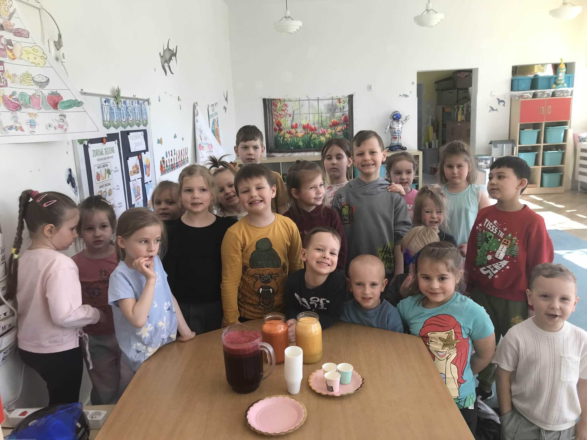 Cała grupa pozuje do wsp&oacute;lnego zdjęcia po zakończeniu zajęć. Przed dziećmi na stoliku znajdują się wykonane soki.