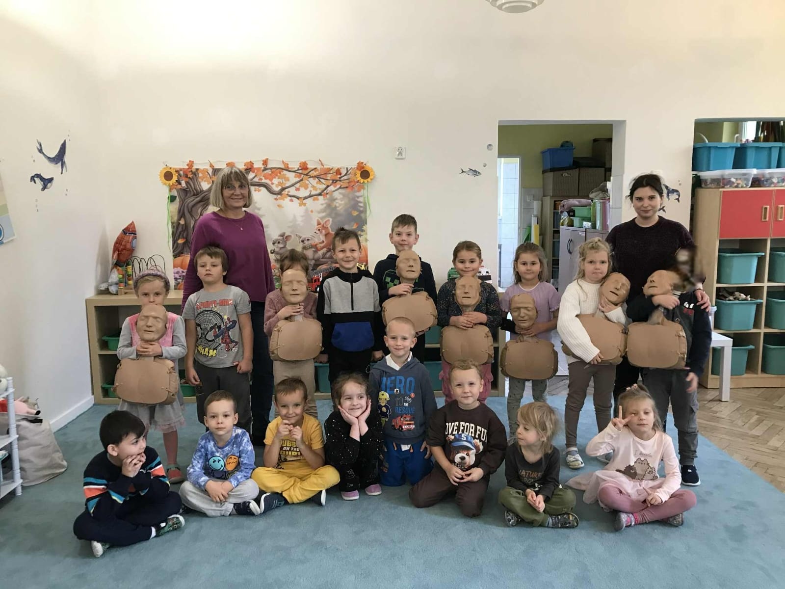 Dzieci i nauczycielki pozują do wspólnego zdjęcia w sali przedszkolnej. Dzieci trzymają fantomy do ćwiczeń RKO. Zdjęcie częściowo anonimizowane.