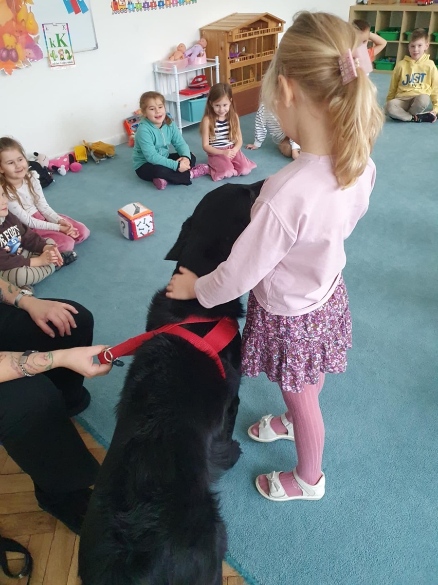 Dzieci siedzą na dywanie. Dziewczynka głaszcze czarnego dużego psa.