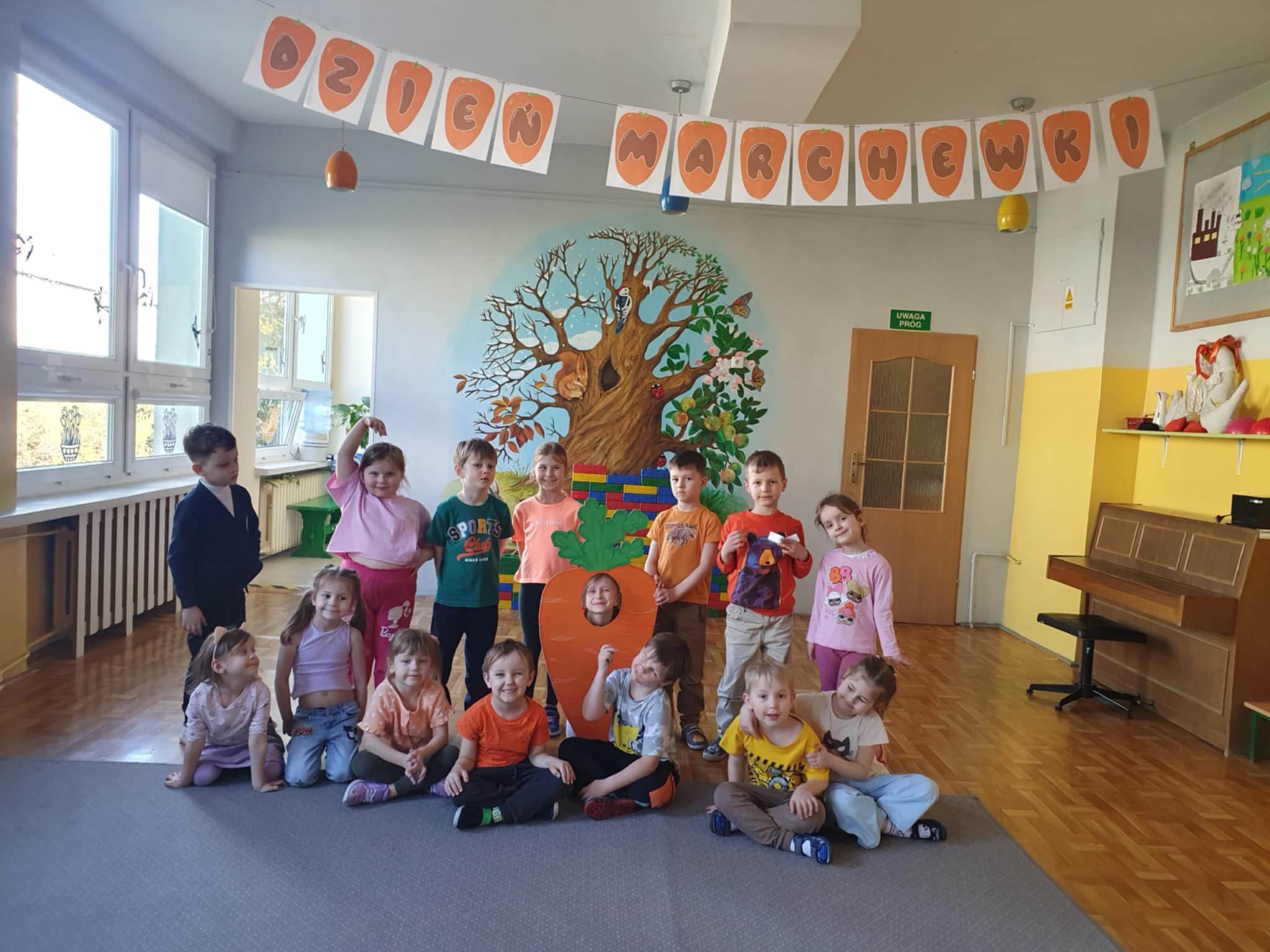 Dzieci pozują do wspólnego zdjęcia w holu przedszkola. Jedno z dzieci w środku kadru trzyma makietę marchewki. W górnej części zdjęcia znajduje się napis "Dzień Marchewki".
