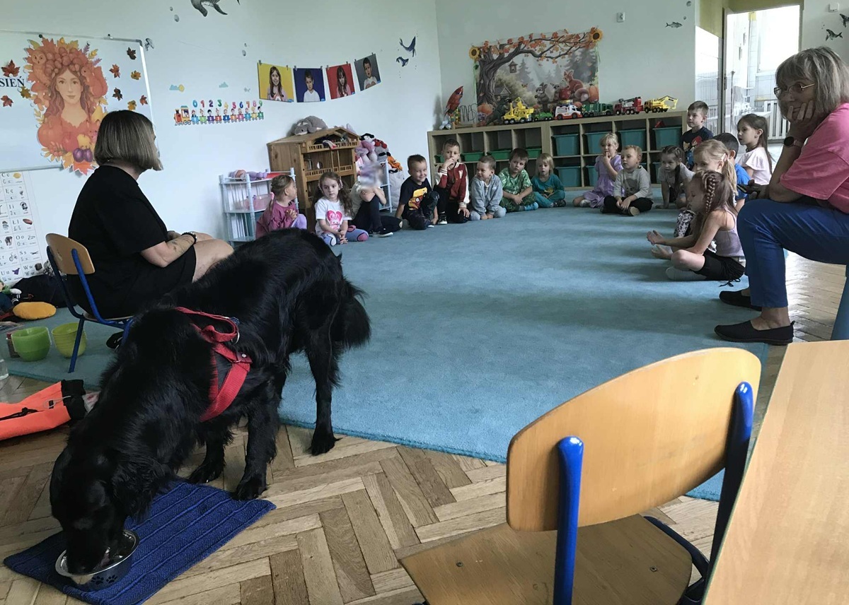 Grupa dzieci siedzi na dywanie w sali i przygląda sie duzemu carnemu psu. Zdjęcie częściowo anonimizowane.