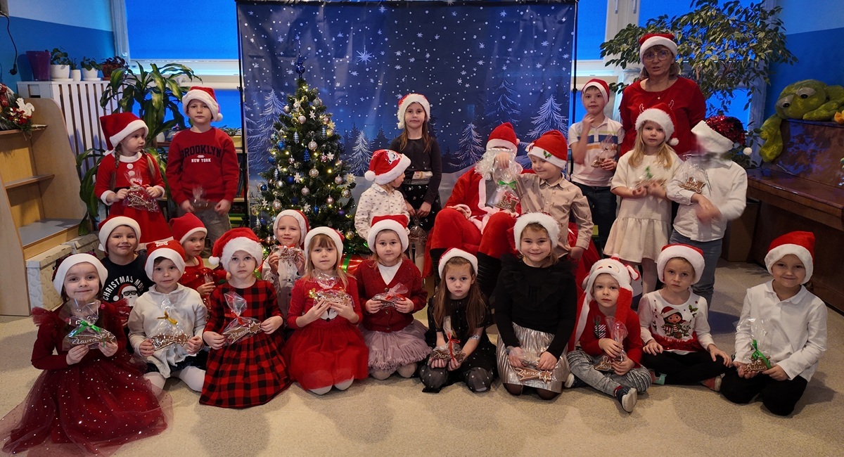 Grupa dzieci i nauczycielka pozują do wsp&oacute;lnego zdjęcia z Mikołajem. Dzieci mają na głowach charakterystyczne czapki. Mikołaj siedzi na fotelu. Zdjęcie częściowo anonimizowane.