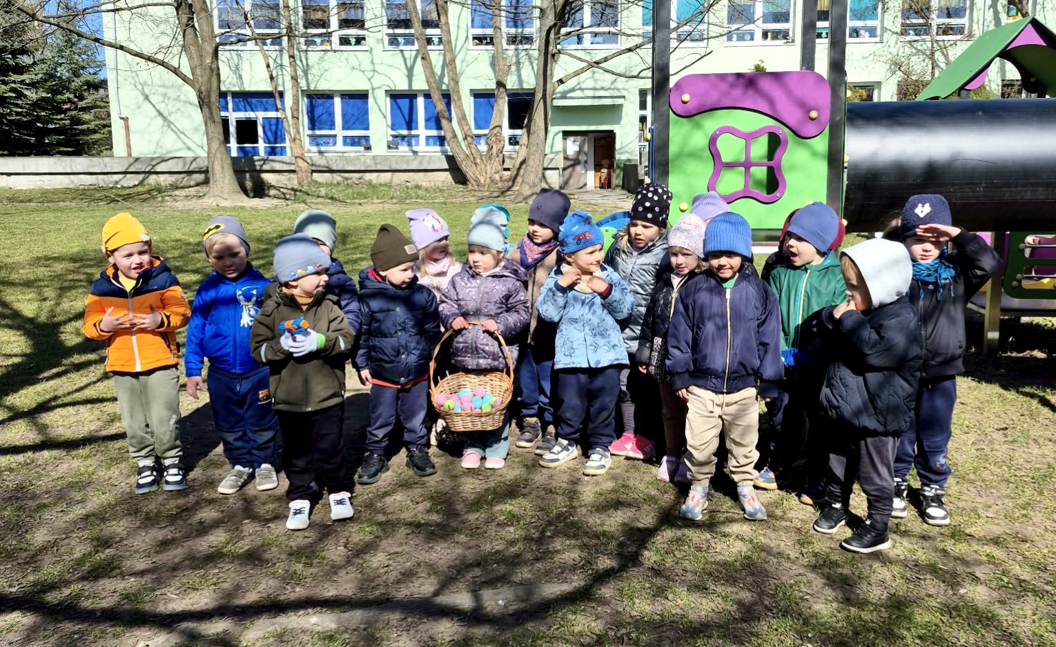 Grupa dzieci pozuje do wsp&oacute;lnego zdjęcia w ogrodzie przedszkolnym na tle zabawki ogrodowej. W tle widać budynek przedszkola. Dzieci trzymają pleciony koszyk wypełniony kolorowymi jajkami.