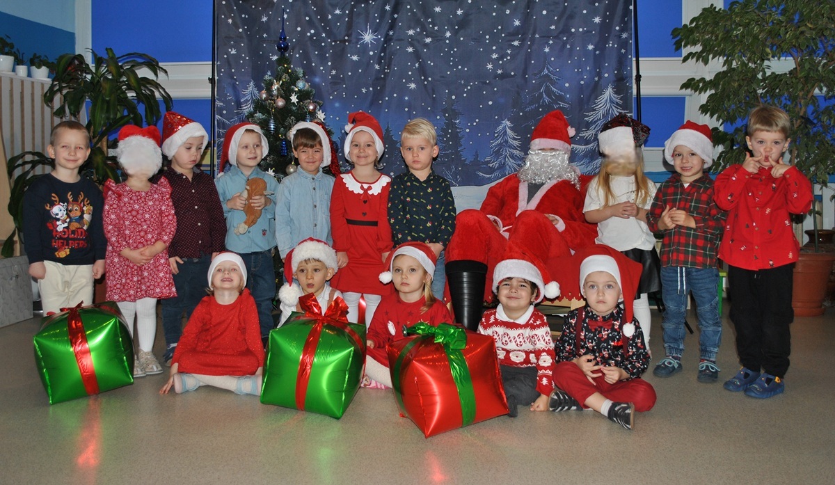 Grupa dzieci pozuje do wsp&oacute;lnego zdjęcia z Mikołajem. Dzieci mają na głowach charakterystyczne czapki. Mikołaj siedzi na fotelu. Zdjęcie częściowo anonimizowane.