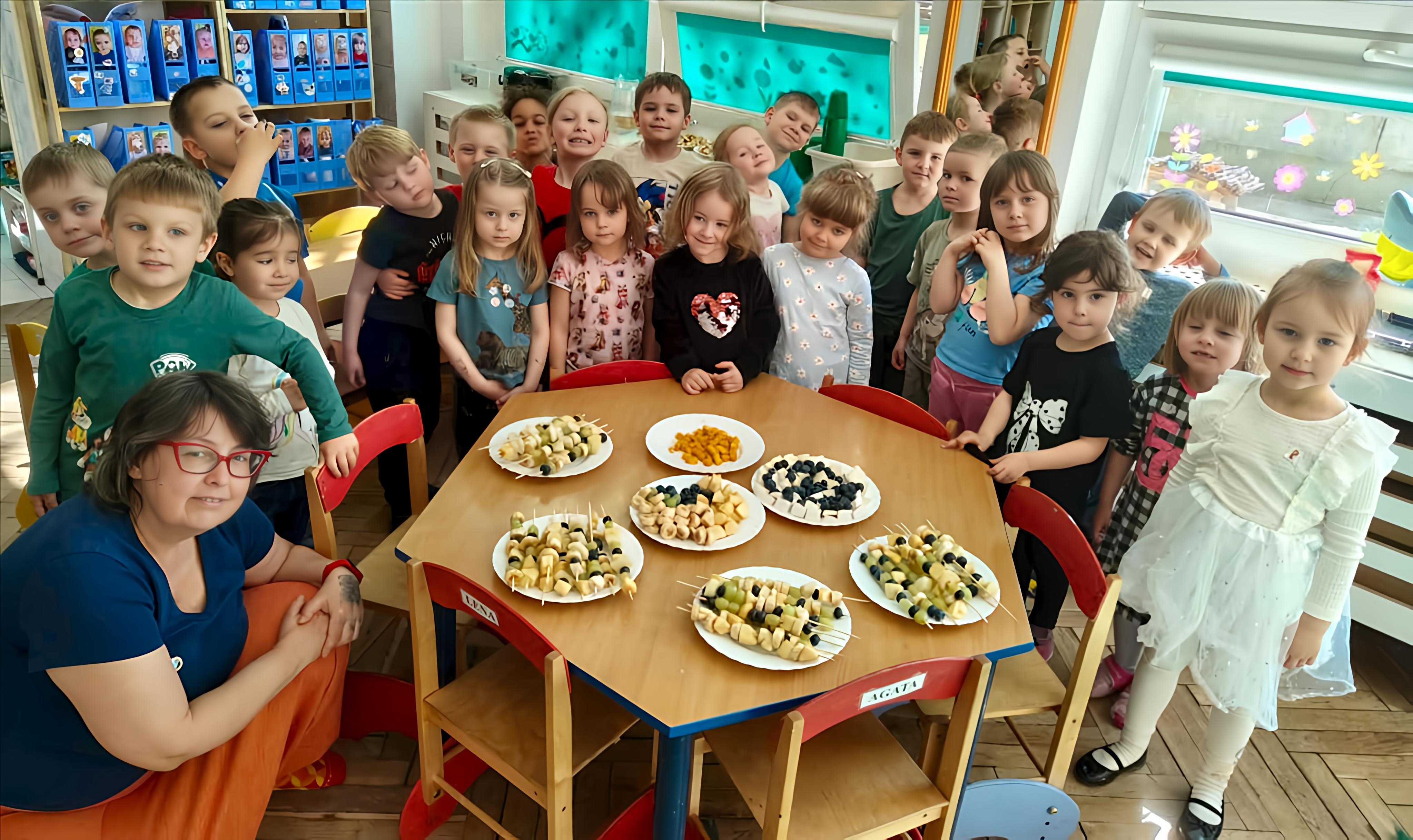 Dzieci i nauczycielka z Grupy 2 pozują do wsp&oacute;lnego zdjęcia po wykonaniu owocowych szaszłyk&oacute;w, kt&oacute;re teraz leżą na talerzach na pierwszym planie zdjęcia.