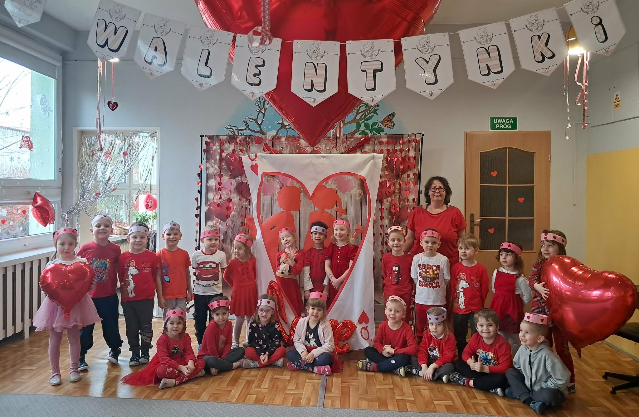 Dzieci i nauczycielka pozują do wsp&oacute;lnego zdjęcia na tle walentynkowych dekoracji.