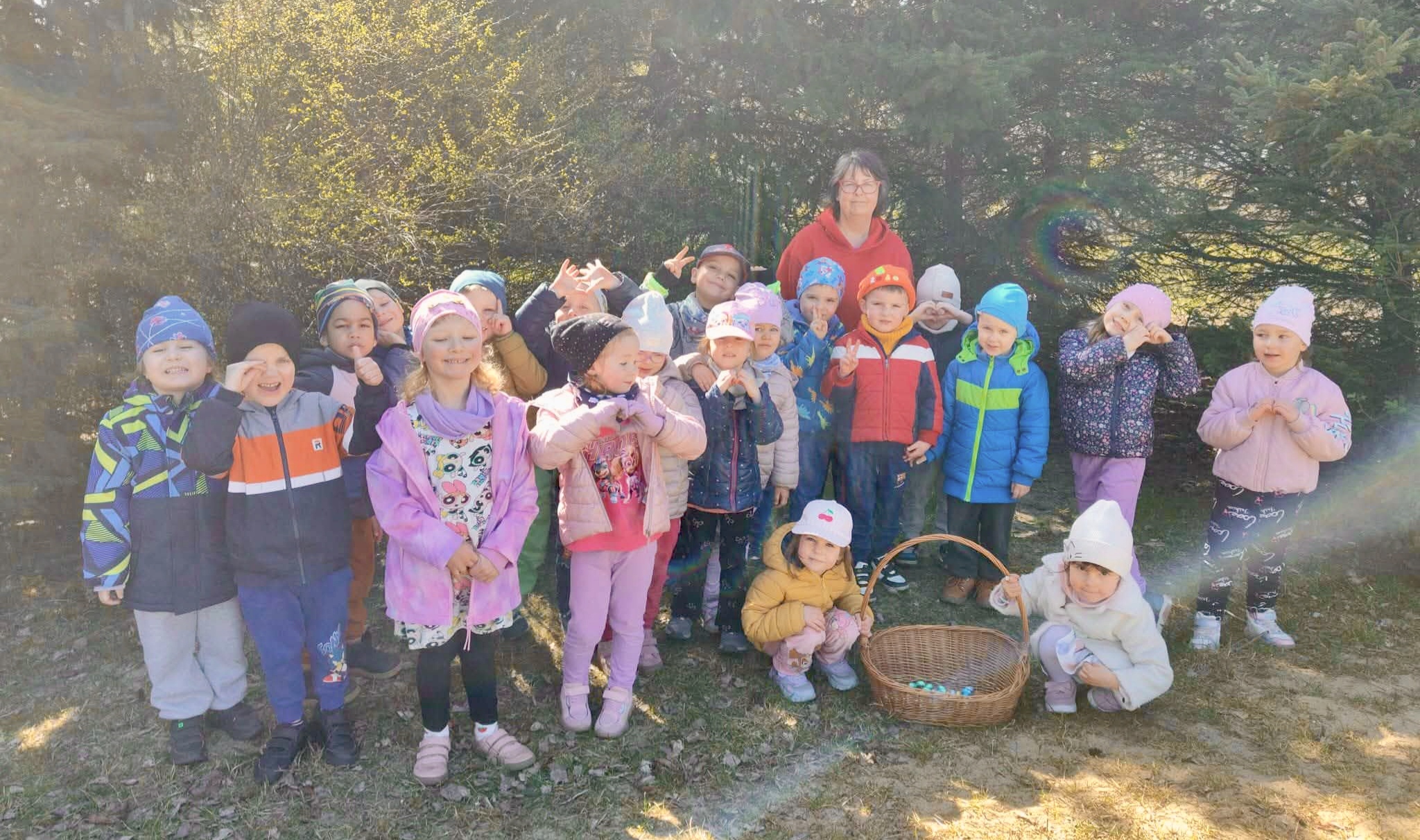 Dzieci i nauczycielka pozują do wspólnego zdjęcia w ogrodzie. Dzieci trzymają pleciony kosz wypełniony czekoladowymi jajkami.