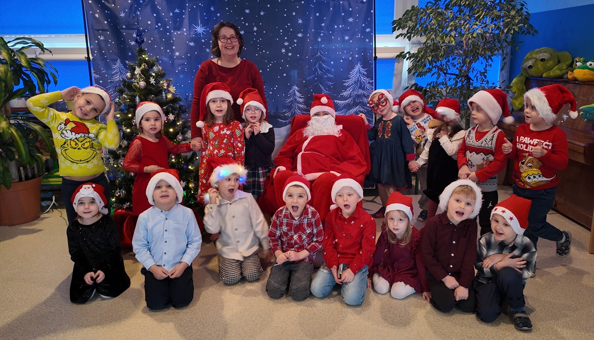 Grupa dzieci i nauczycielka pozują do wsp&oacute;lnego zdjęcia z Mikołajem. Dzieci mają na głowach charakterystyczne czapki. Mikołaj siedzi na fotelu. Zdjęcie częściowo anonimizowane.