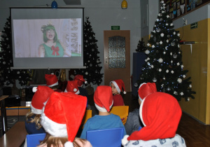 Dzieci oglądają na dużym ekranie nagranie występu "Pani Choinki". Dzieci mają na głowach czapki Mikołaja. Ujęcie 1