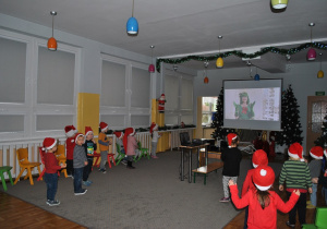 Dzieci w mikołajowych czapkach tańczą tak jak pokazuje Pani Choinka. Ujęcie 1