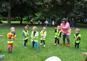 Dzieci razem z nauczycielką tańczą w ogrodzie. Dzieci ubrane są w kamizelki odblaskowe.