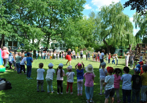 Duża grupa dzieci tworzy koło dookoła konferansjerki. Ujęcie spod drzew.
