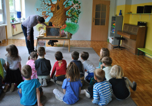 Pan pszczelarz opowiada dzieciom o pszczołach i pokazuje rekwizyty. Dzieci siedzą na dywanie.
