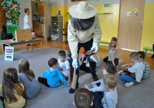 Pan pszczelarz w stroju ochronnym pokazuje dzieciom rekwizyty.