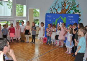Dzieci stoją w półkolu ustawione na scenie i śpiewają piosenkę.