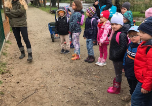 Dzieci stoją przed wybiegiem dla zwierząt. Za dziećmi stoi nauczyciel. Z boku stoi przewodniczka i opowiada o zwierzętach.