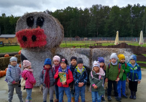 Dzieci pozują do wspólnego zdjęcia. W tle posąg zbudowany z pomalowanych belek słomy.