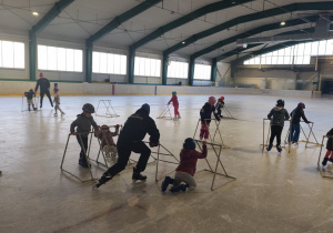 Dzieci w czasie nauki jazdy na łyżwach. Na zdjęciu widoczni są trenerzy. Dzieci utrzymują równowagę korzystając ze specjalnie przygotowanych sanek.