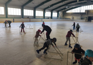 Dzieci w czasie nauki jazdy na łyżwach. Na zdjęciu widoczni są trenerzy. Dzieci utrzymują równowagę korzystając ze specjalnie przygotowanych sanek.