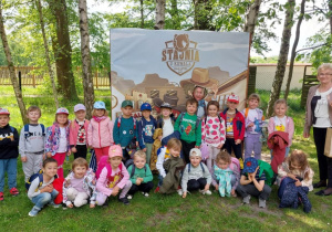 Dzieci pozują do pamiątkowego wspólnego zdjęcia w czasie wycieczki do Nieborowa. Grupa dzieci stoi przed banerem reklamowym z napisem "Stajnia u Kowala".