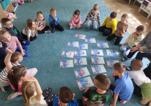 Dzieci siedzą w kole na dywanie i oglądają ilustracje z morskimi stworzeniami. Razem z dziećmi siedzi nauczycielka. Zdjęcie częściowo anonimizowane.