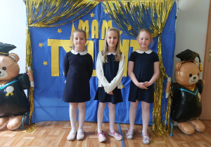 Troje dzieci pozuje do wspólnego zdjęcia na tle napisu "Mam talent". Dzieci ubrane w galowe sukienki.