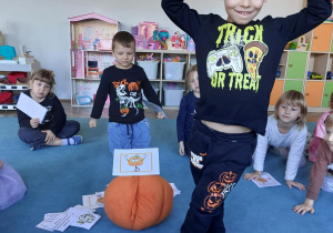 Dzień dyni w grupie 3. Dzieci siedzą na dywanie, a jedno z nich stoi podnosząc ręce.