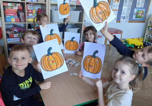 Dzień dyni w grupie 3. Dzieci siedzą przy stoliku i prezentują prace plastyczne.