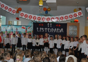 Przedstawienie z okazji Dnia Niepodległości. Dzieci z grupy 4 i 5 stoją razem na scenie przed widownią.