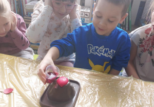 Dzieci siedzą przy stoliku. Chłopiec trzyma kubek z czerwoną cieczą.