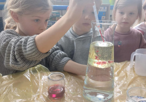 Dzieci siedzą przy stoliku. Dziewczynka trzyma flakon z płynem i pipetę z czerwonym płynem.