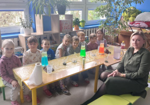 Dzieci i nauczycielka siedzą przy stoliku, na którym stoi wyposażenie z zajęć chemicznych.