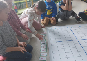 Dzieci siedzą dookoła planszy do kodowania i oglądają kolorowe karty.