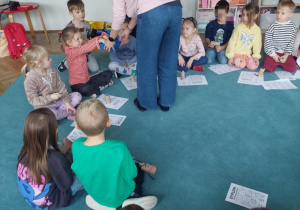 Dzieci siedzą w kole na dywanie. Przed dziećmi leżą karty pracy. Nauczycielka stoi przy jednym z dzieci i podaje mu pudełko, a dziecko coś losuje.