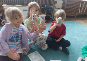 Troje dzieci prezentuje samodzielnie wykonane kotki z tekturek po papierze toaletowym.