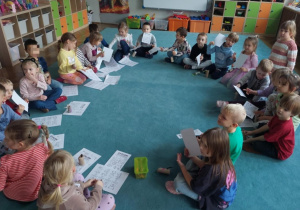 Dzieci siedzą w kole na dywanie, a przed nimi leżą karty pracy.