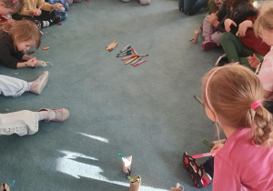 Dzieci i nauczycielka siedzą na dywanie. Przed dziećmi stoją elementy i materiały do wykonania pracy plastycznej.