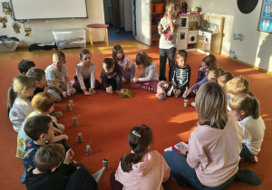 Dzieci i nauczycielka siedzą na dywanie. Przed dziećmi stoją wykonane prace - kotki z tekturowych rolek po papierze toaletowym. Ujęcie 1
